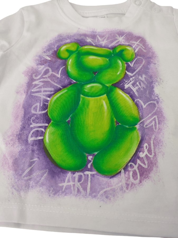 La camiseta arte infantil refleja encanto y color en cada pincelada pintando este osito verde sobre fondo morado para bebé de 12 meses